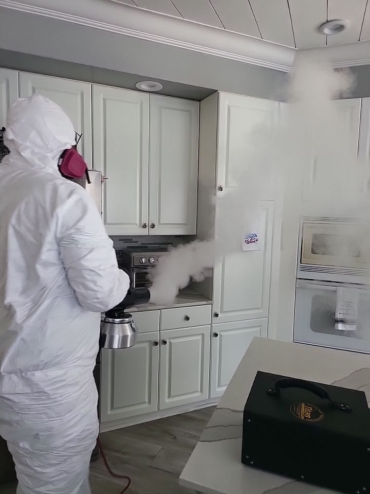 Technician in full PPE thermal fogging kitchen cabinets with air scrubber on floor